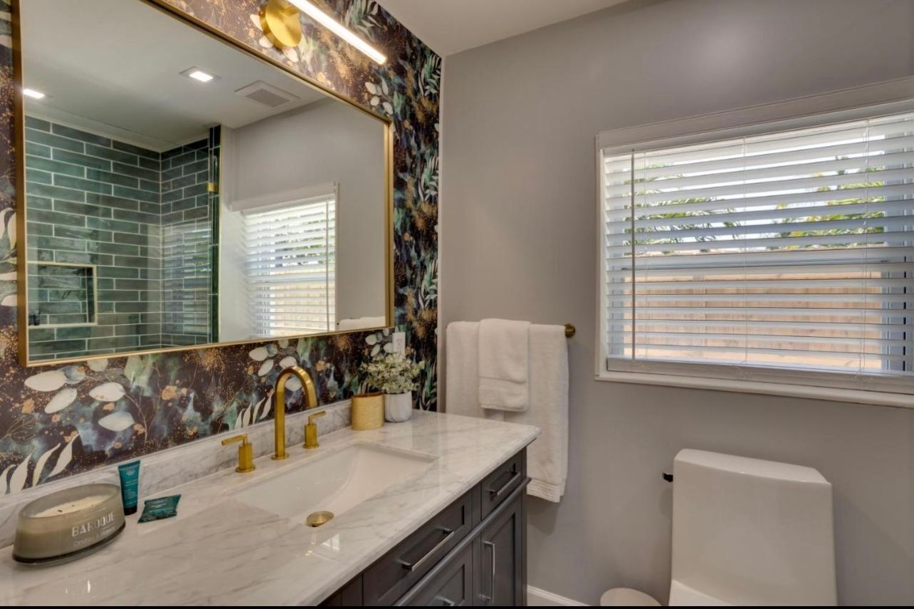 Bathroom with marble vanity, floral wallpaper, gold fixtures, and a window with blinds.