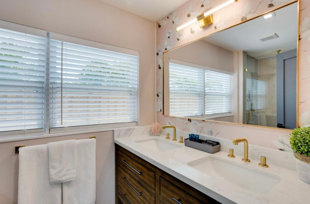 Bathroom with double sinks, gold fixtures, pink walls, and white blinds.