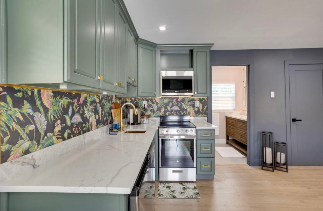 Green kitchen with floral backsplash, stainless steel appliances, and hardwood floors.