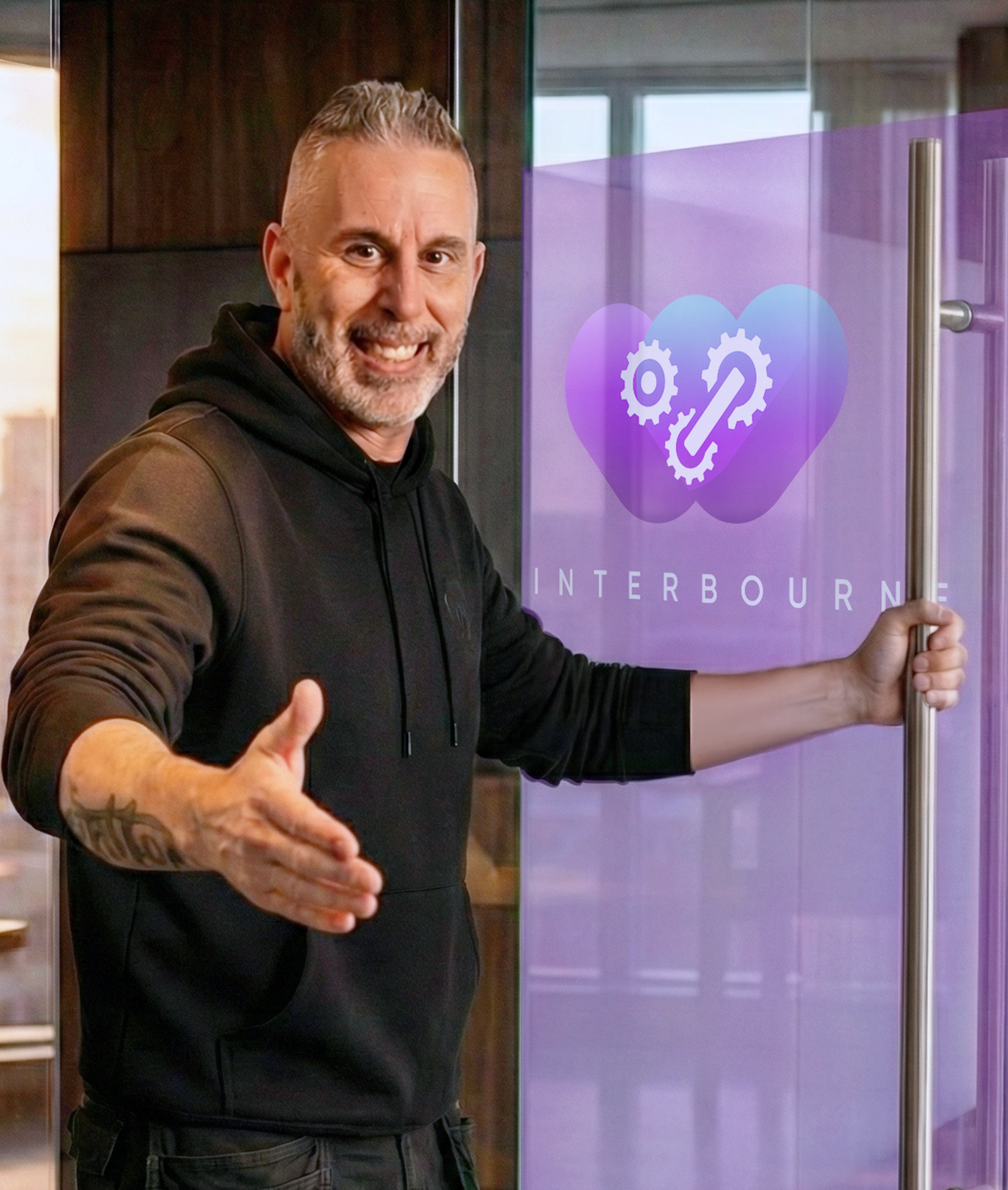 Man in black hoodie smiles, extends hand, stands near a glass door with a purple logo.