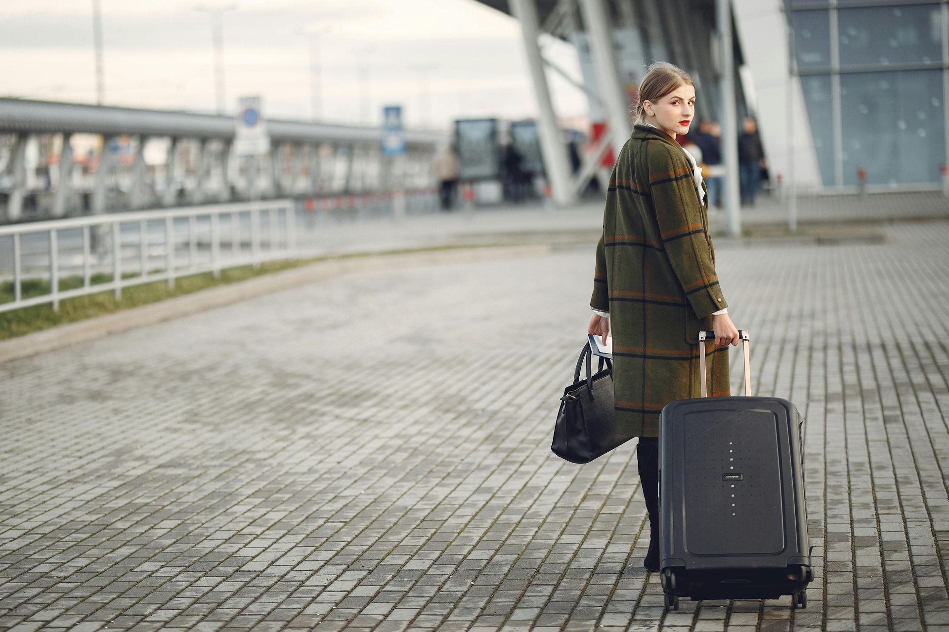 Woman with luggage outside an airport terminal. She wears a long green coat and looks back.
