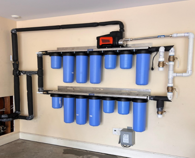 Water filtration system with blue filter housings and black pipes mounted on a beige wall in a garage.