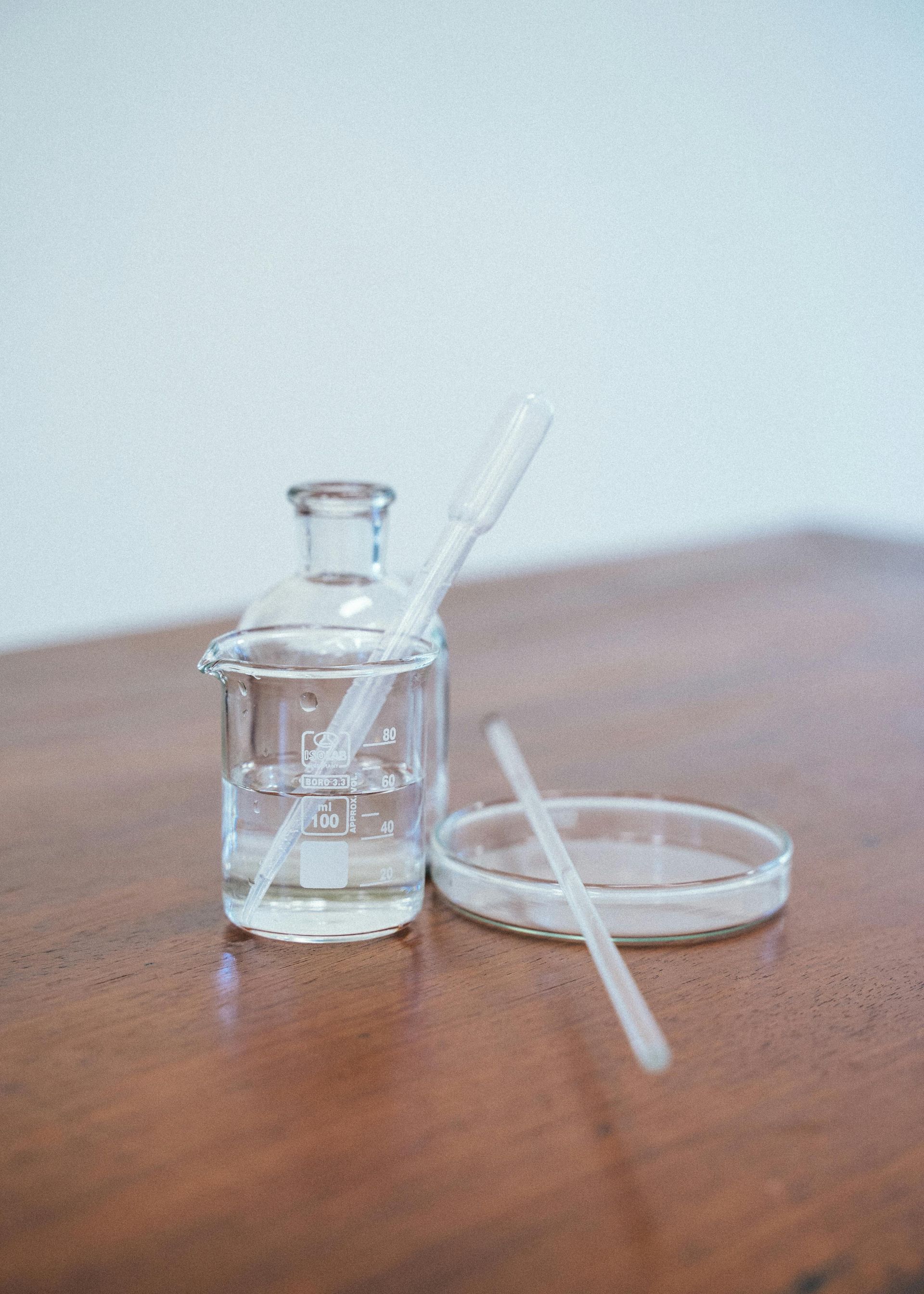 Laboratory glassware: a glass bottle with liquid, a petri dish, and pipettes on a wooden surface.