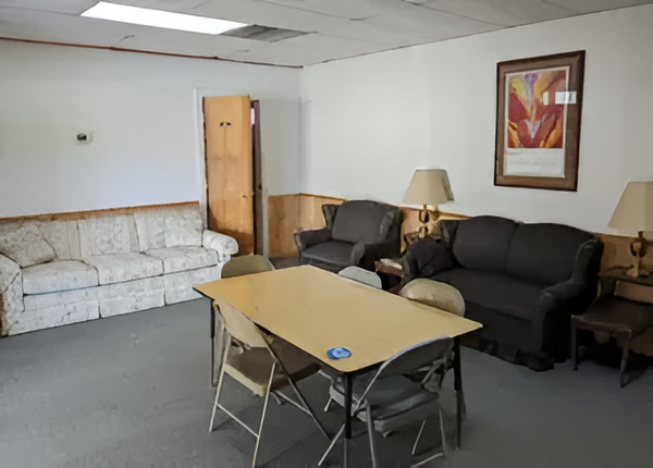 A furnished community room with a table and folding chairs, a sofa, two armchairs, and framed artwork on the wall.