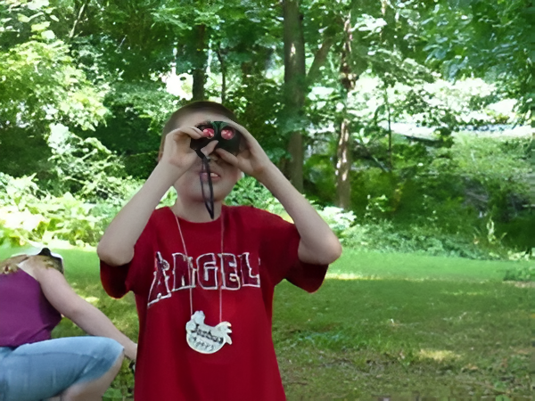 Boy in red shirt using binoculars, looking up in a green, wooded area.
