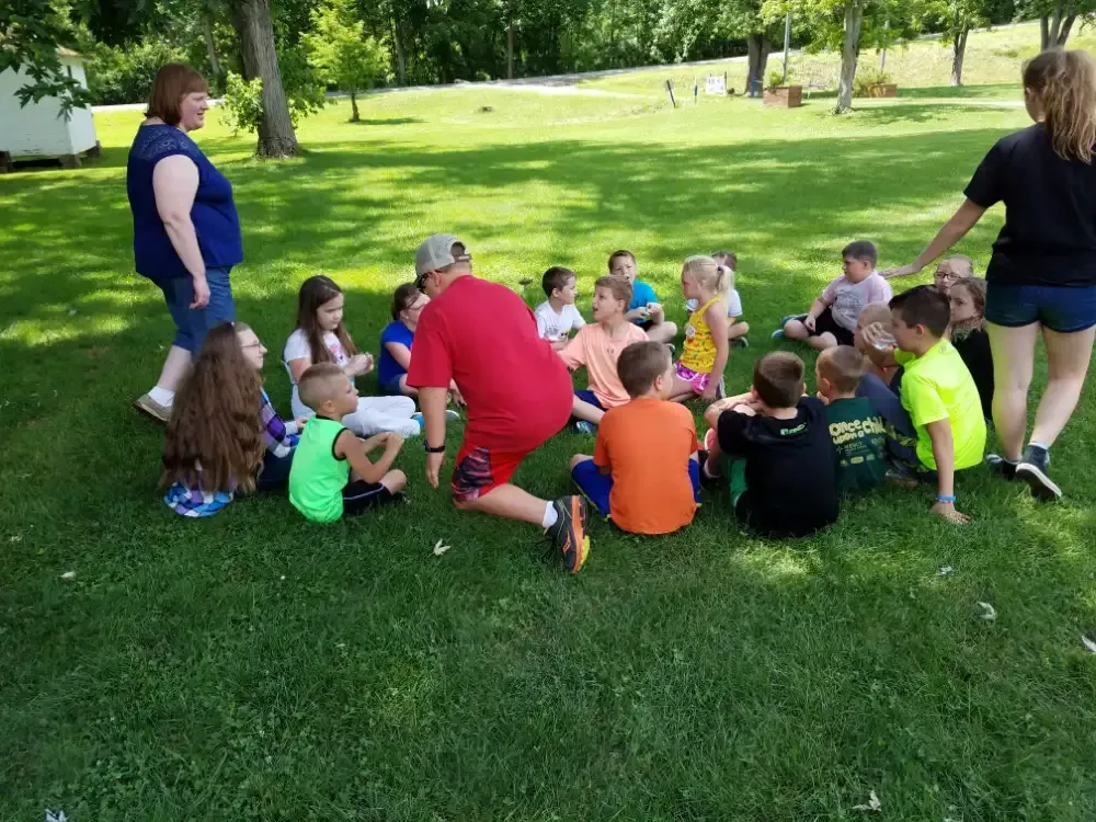 A group of children and adults are sitting in a circle on a grassy lawn, possibly for a lesson or activity.