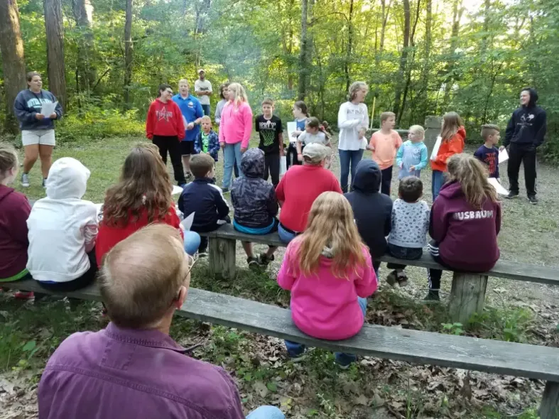 Children and adults seated on benches in a wooded area, some facing each other, possibly at a camp.