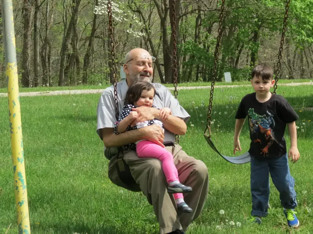A grandfather swings with a toddler on a swing set, with a boy watching. Outdoors, green grass.