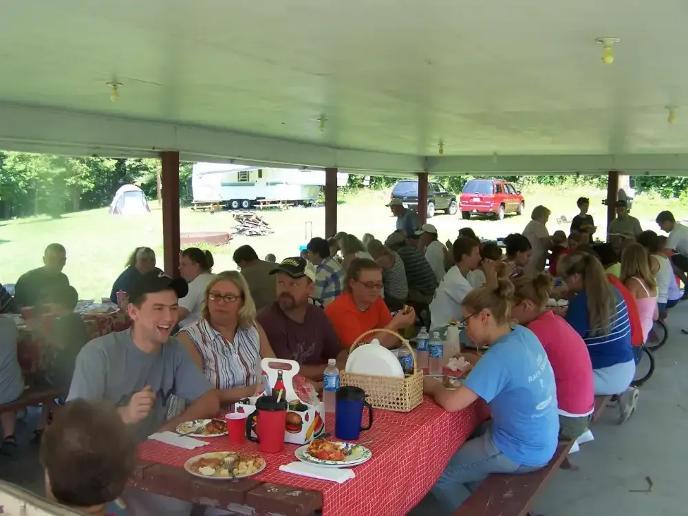 People gathered at a picnic table under a shelter, eating and socializing. Red checkered tablecloth.