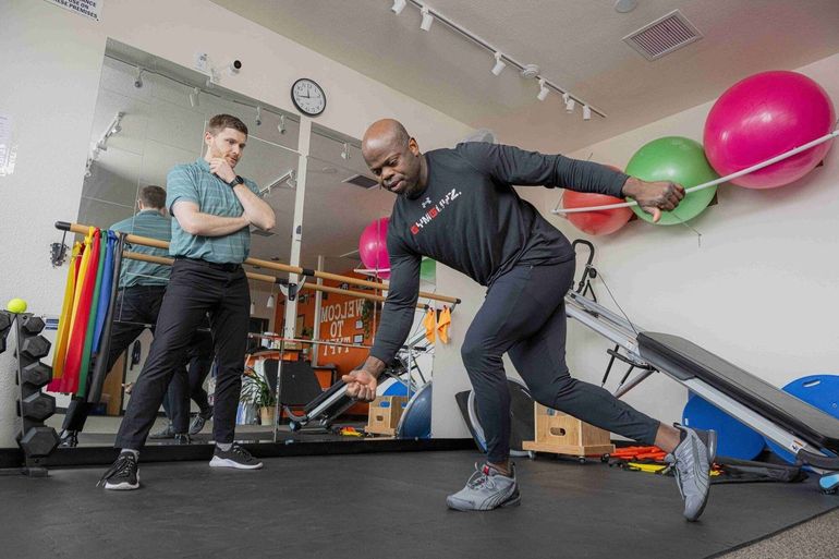 Two people exercising in a gym, one balancing in a forward lunge beside exercise balls.