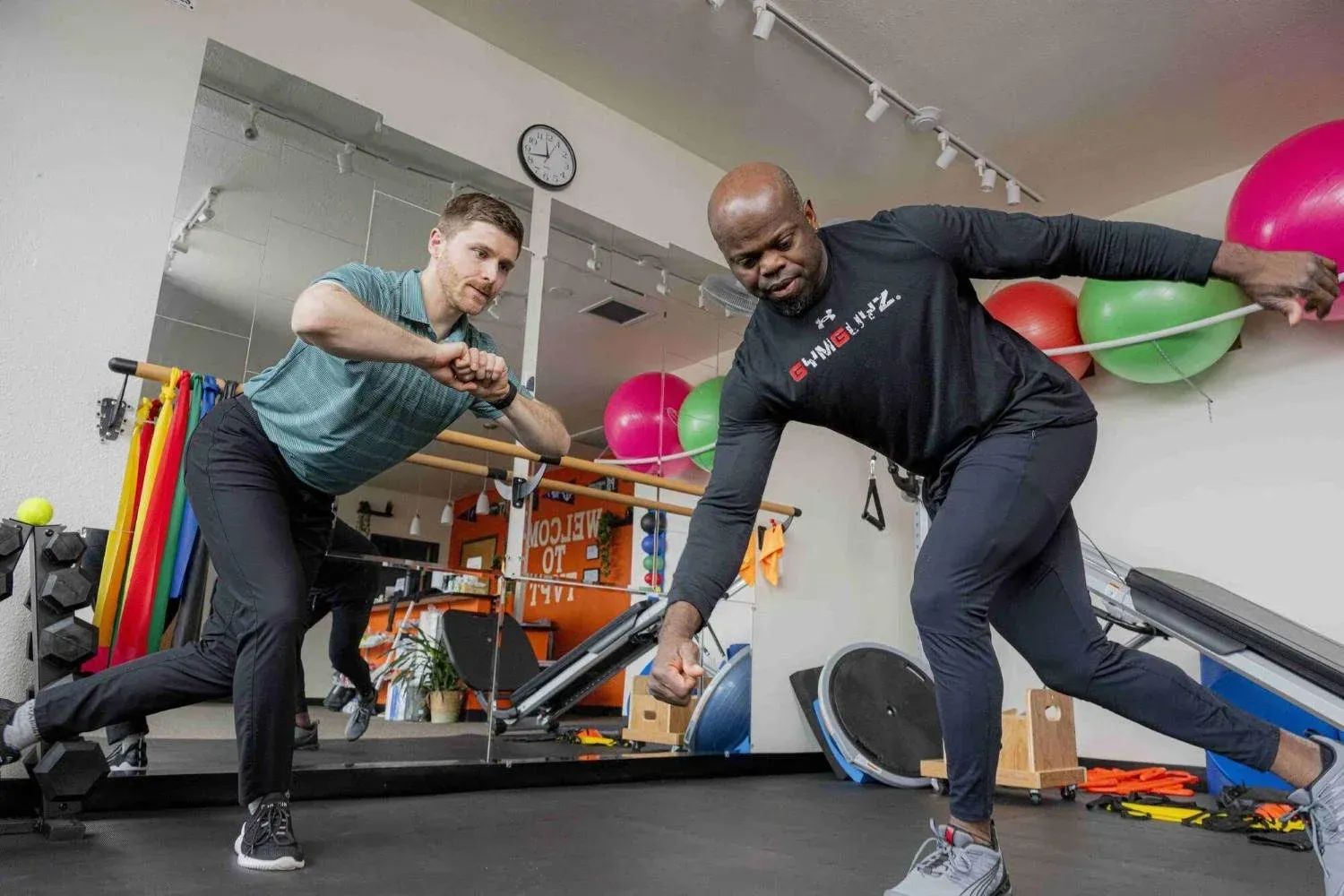 Two people exercising with resistance bands in a gym, surrounded by exercise balls and equipment.