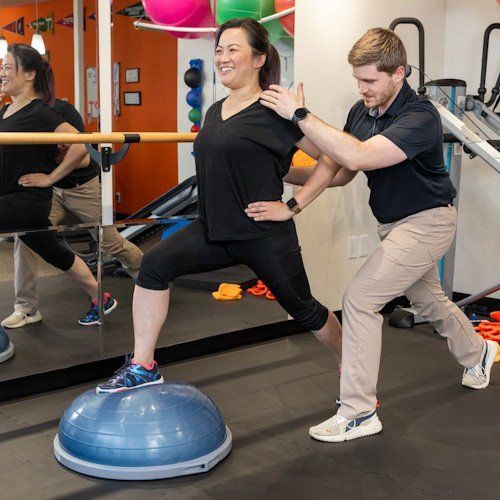 Two people exercising in a gym, one balancing on a blue BOSU ball while the other spots her.