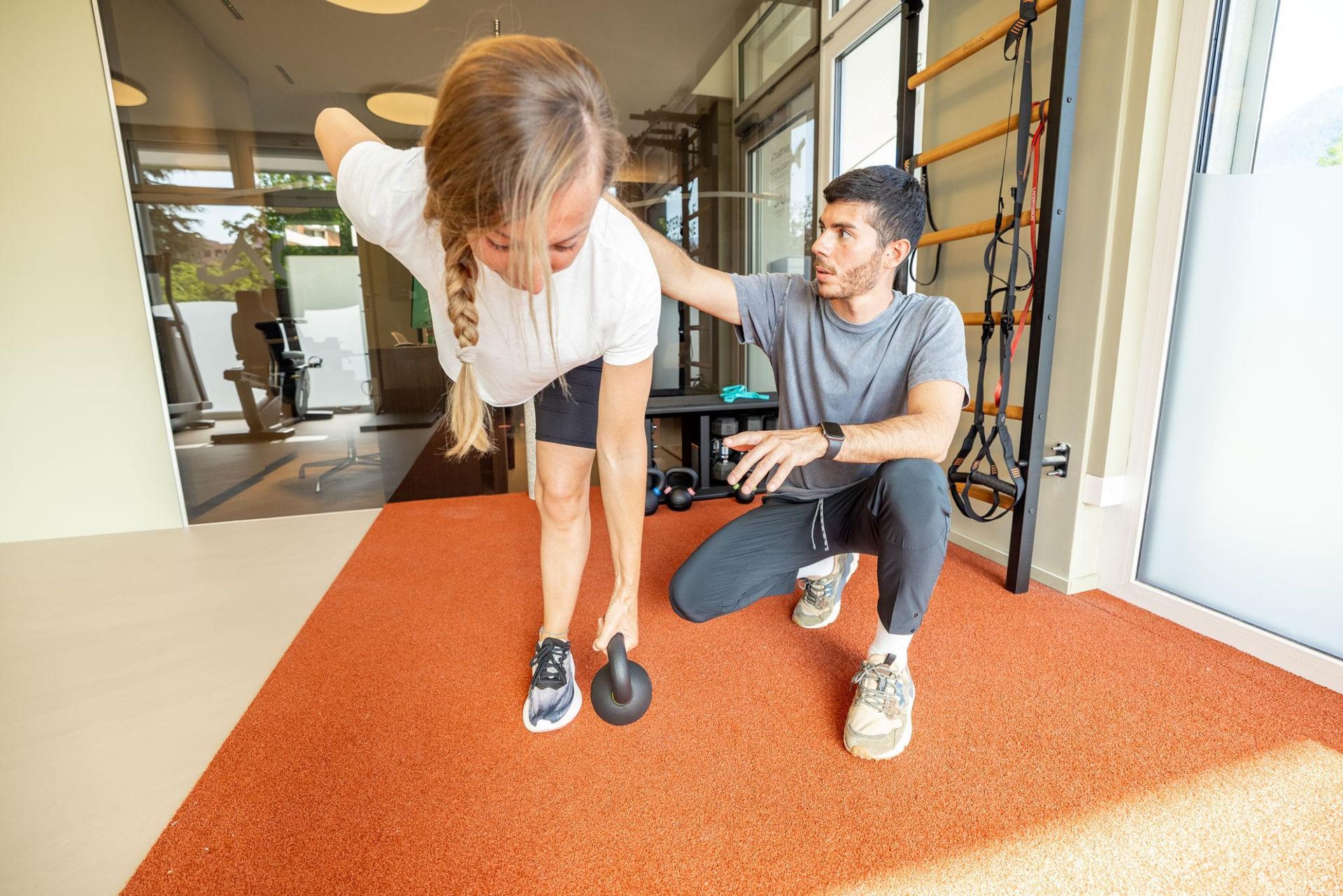 Two people exercising on an orange carpet in a bright indoor gym, one assisting the other with balance.