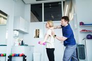 Two people in a bright kitchen, one adjusting the other's shirt collar near a sink and cabinets