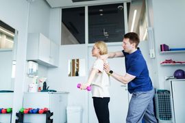 Two people in a bright room, one helping the other adjust clothing near a mirror.