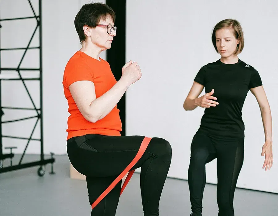 Two people exercising with a resistance band in a studio, one in orange demonstrating a raised knee.