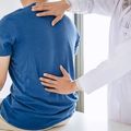 Doctor examining a patient’s back in a clinic, with one hand on the shoulder and one on the lower back.