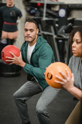 People doing medicine ball squats in a gym, holding red and orange balls.