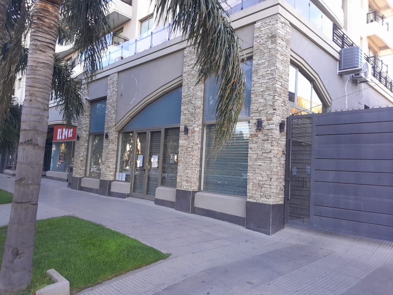Fachada con columnas de piedra, ventanas arqueadas y puerta azul. Palmeras bordean la acera.