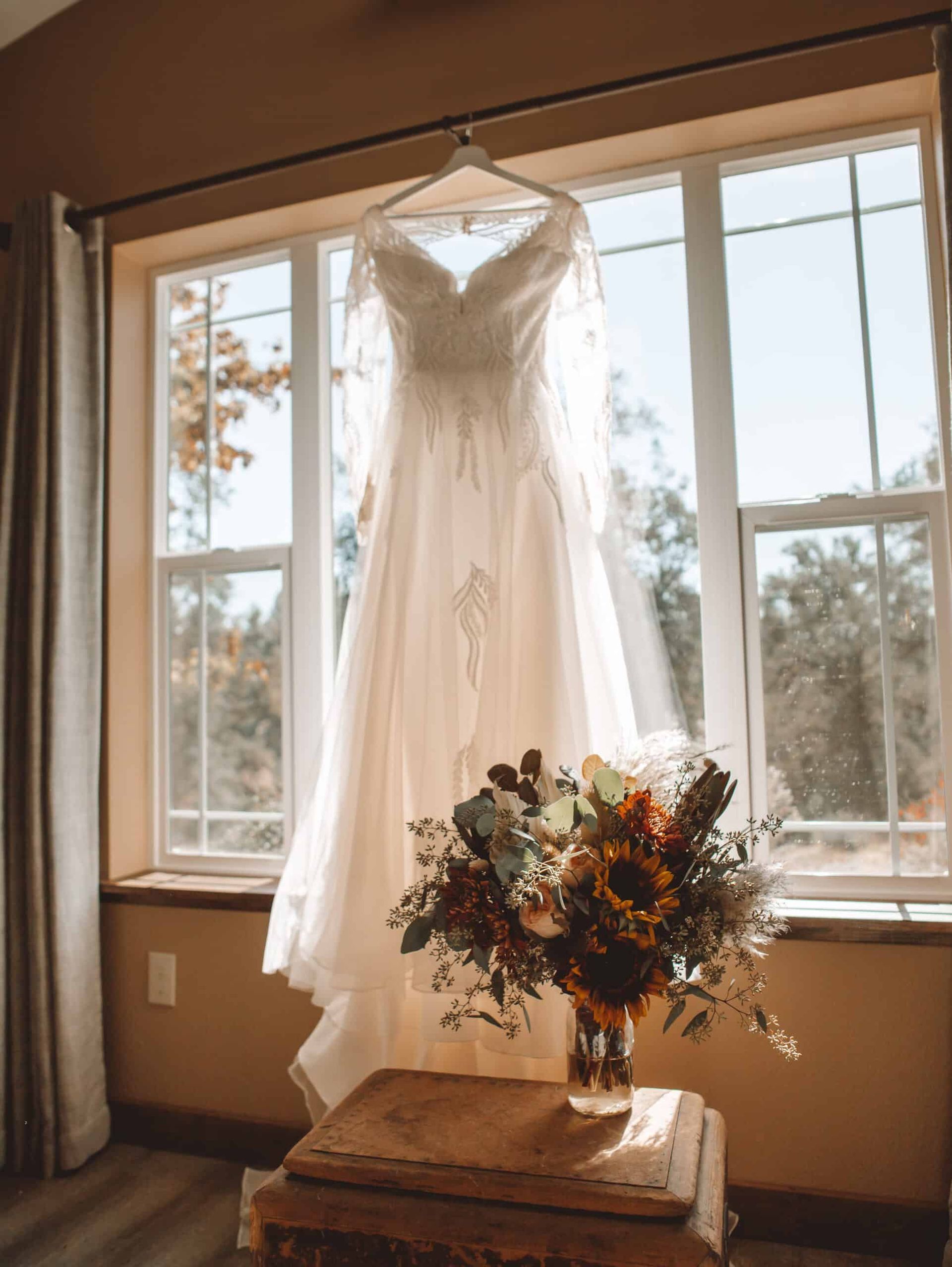 A wedding dress is hanging in a window next to a vase of flowers.