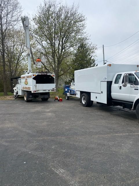 Two white utility trucks on asphalt. One has a raised bucket. Trees and sky in background.