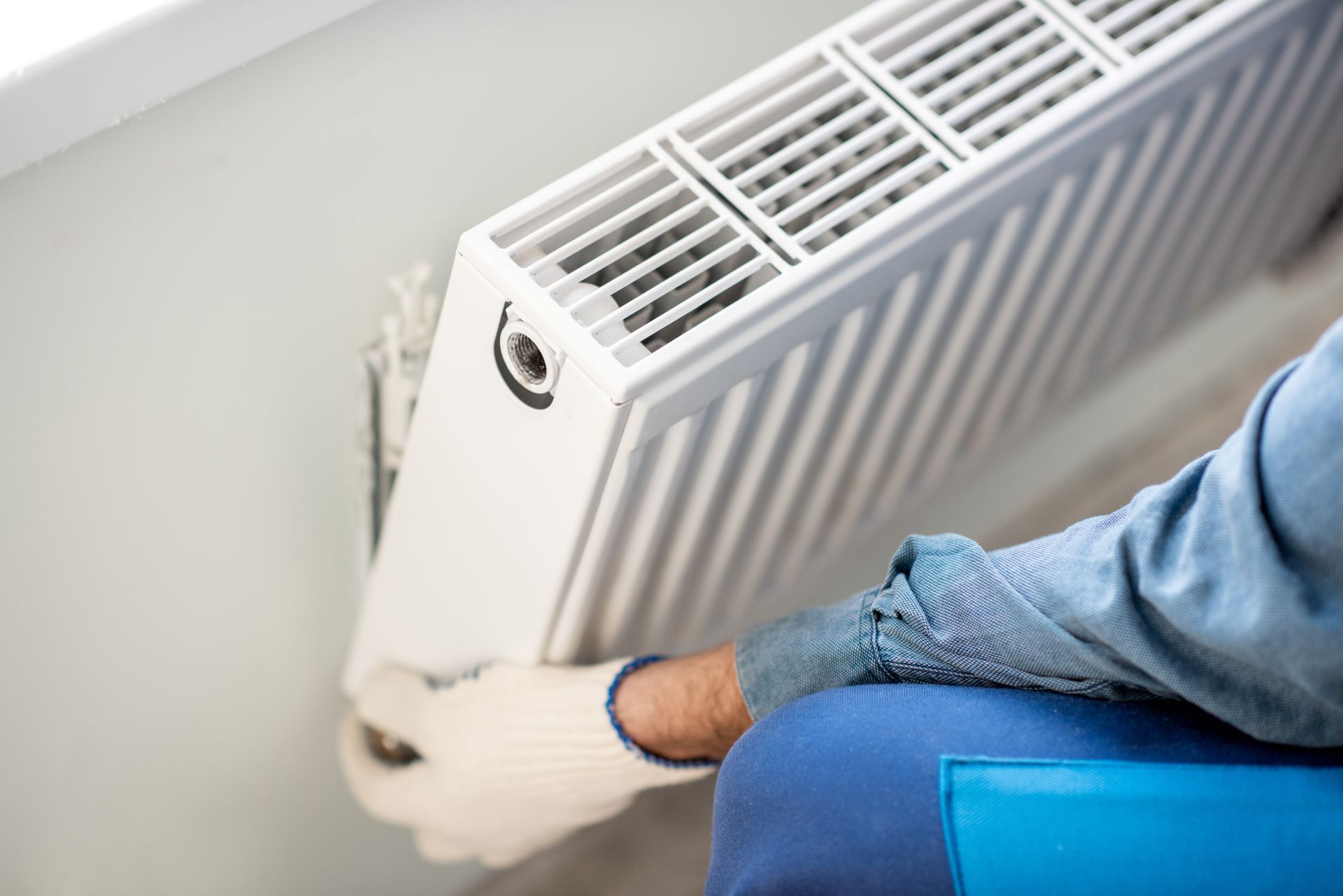 Person adjusting a white radiator with a wrench, wearing a blue sweater in a bright room