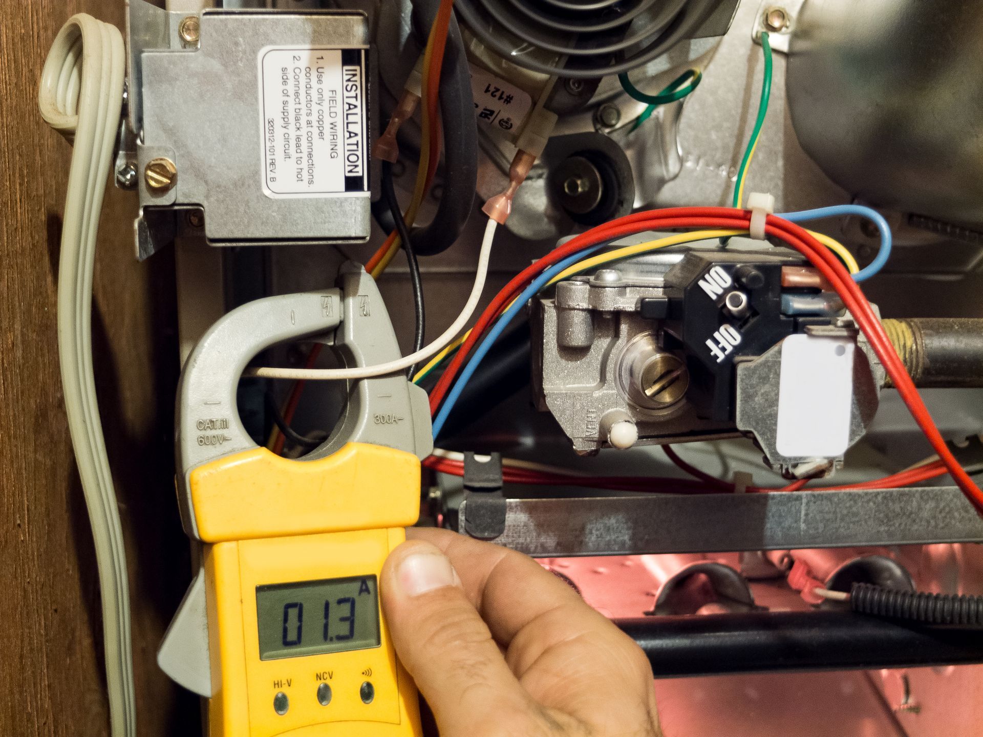 Hand holding a yellow clamp meter measuring a wire inside an open electrical appliance panel