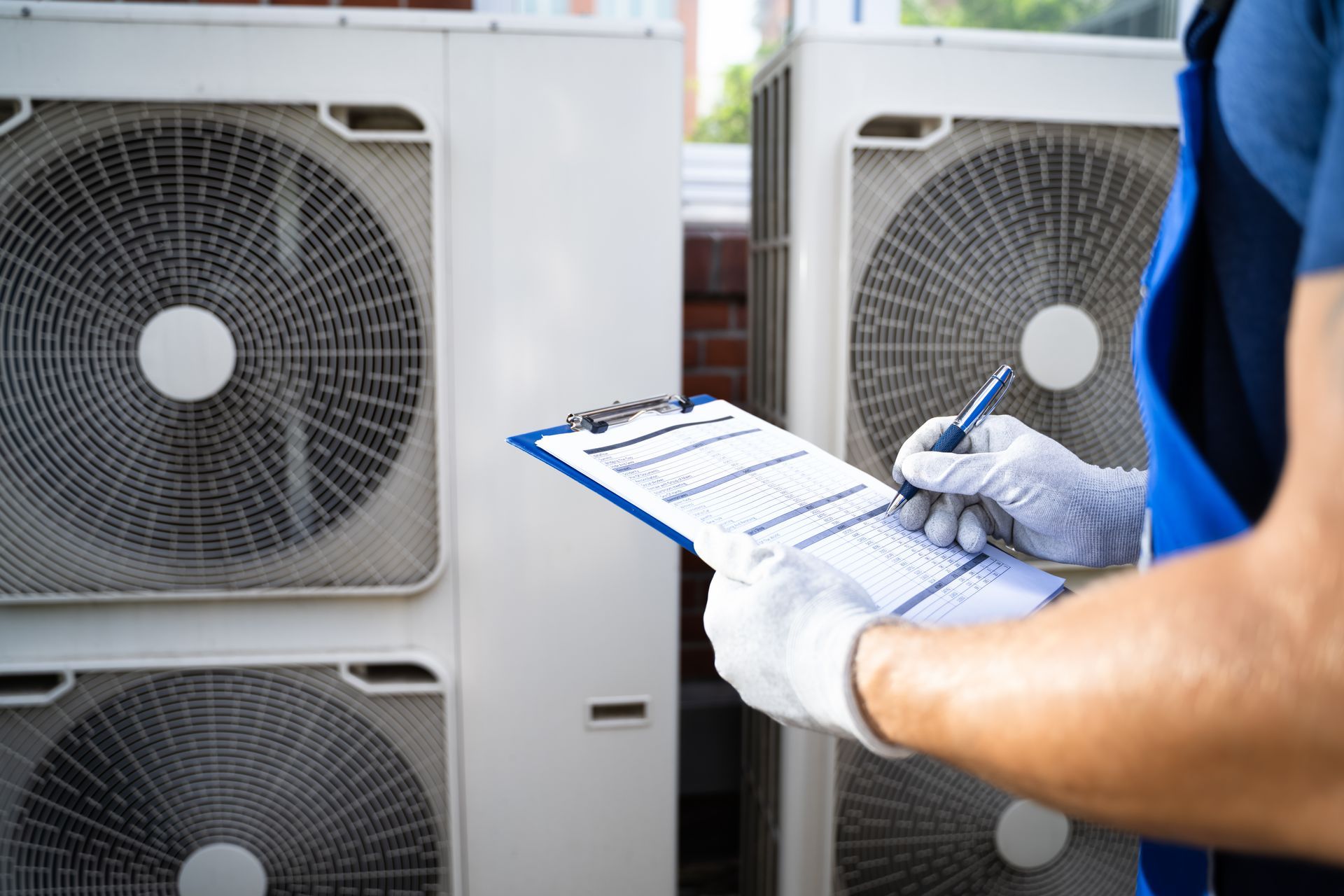 Technician in blue shirt inspecting outdoor air conditioning units with a clipboard and gloves