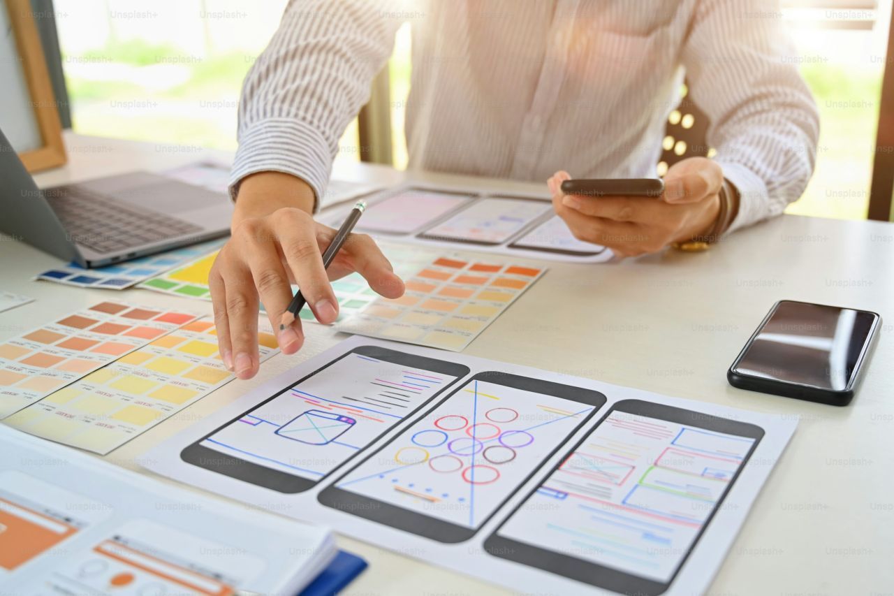 Person designing a mobile app, using a phone, paper mockups, and color swatches.