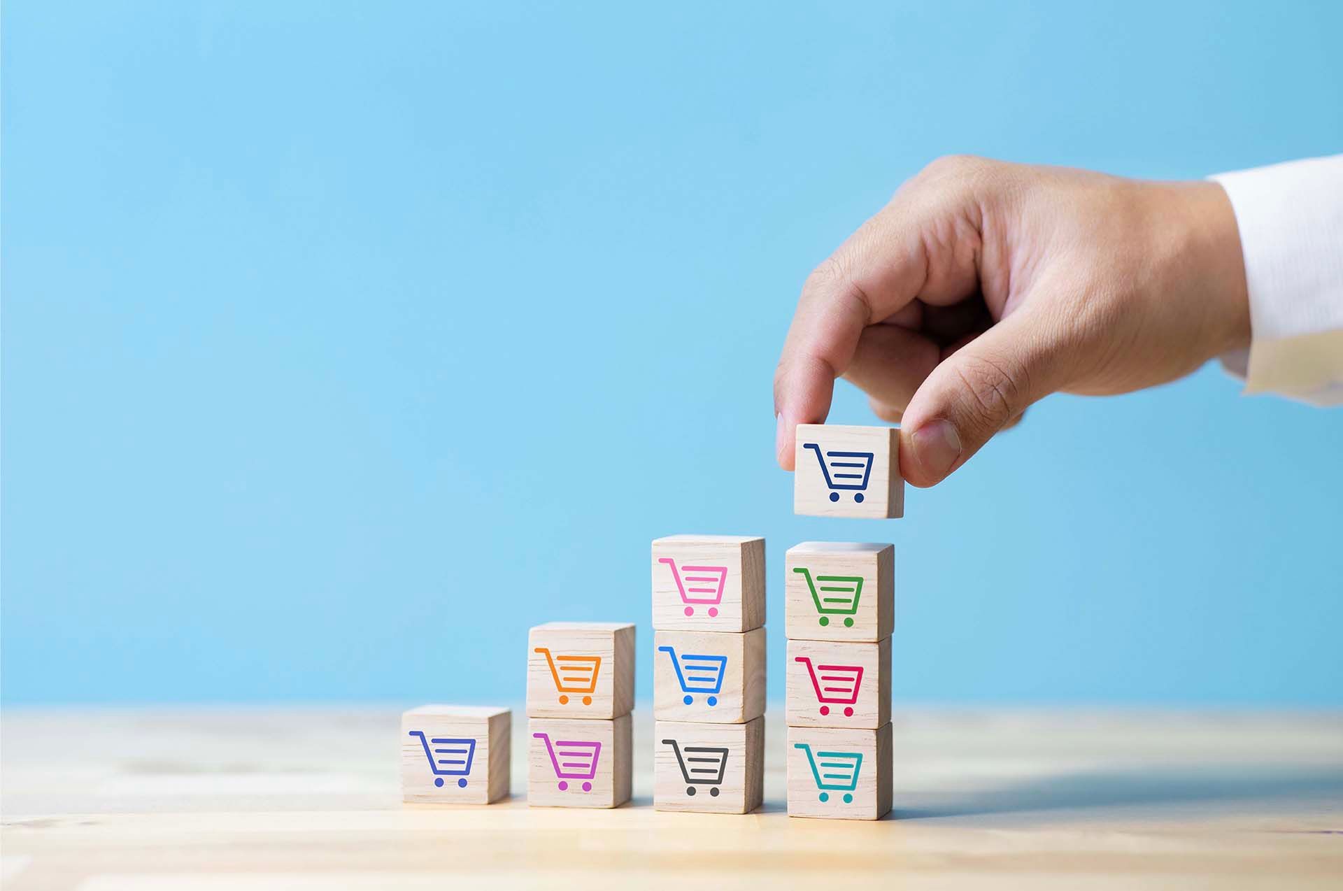 Hand stacking blocks with shopping cart icons, forming a rising bar graph, on a light blue background.