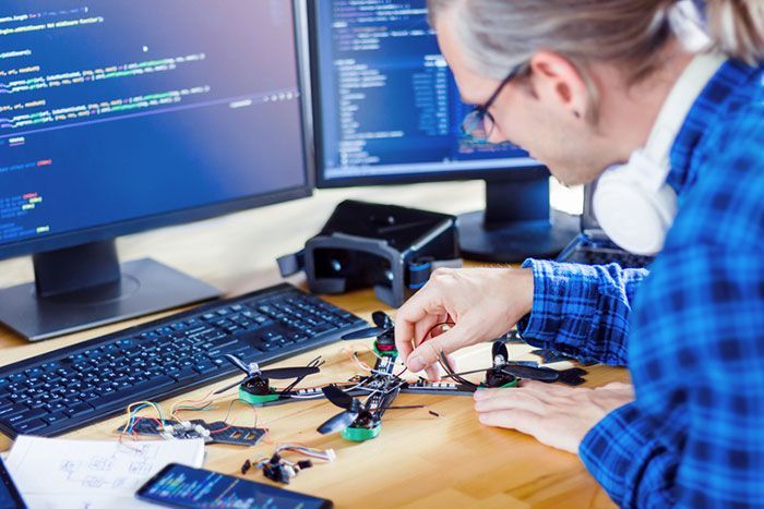 Man working on drone components at a desk with computer monitors displaying code and a VR headset.