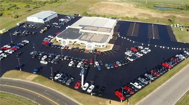 Aerial view of a car dealership with many cars parked on a black asphalt lot, beige building, and green grass.