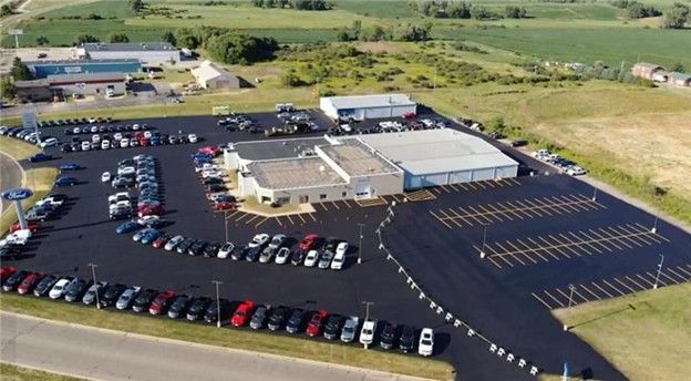 Aerial view of a car dealership, showcasing rows of vehicles on a large paved lot next to buildings and fields.