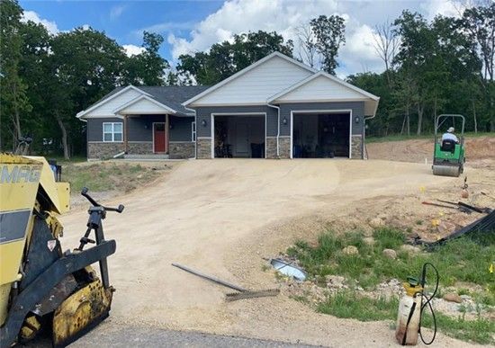 House under construction with two-car garage; workers compacting gravel driveway on a sunny day.