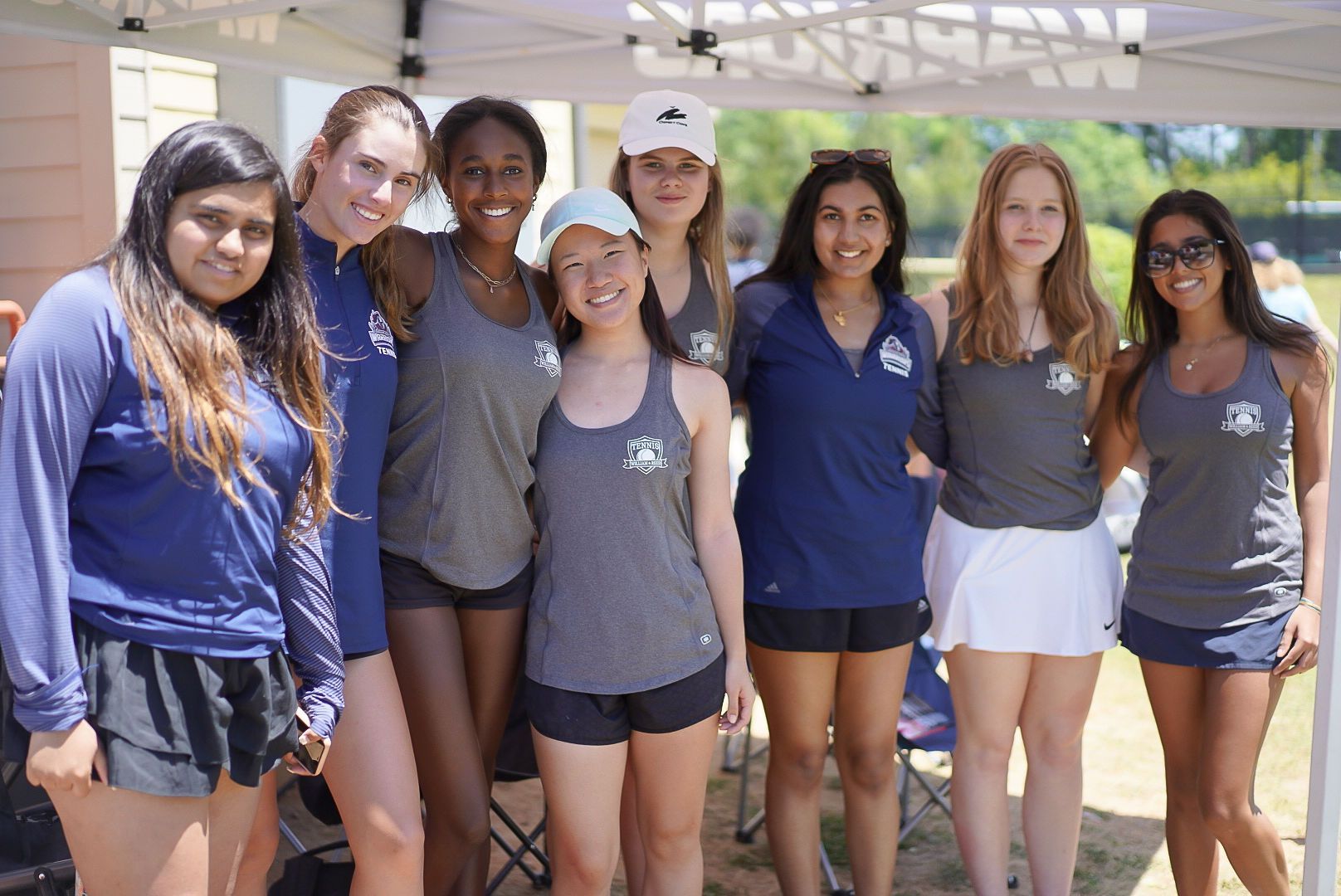 Group of eight people smiling outdoors in matching blue shirts, standing together in bright sunlight.