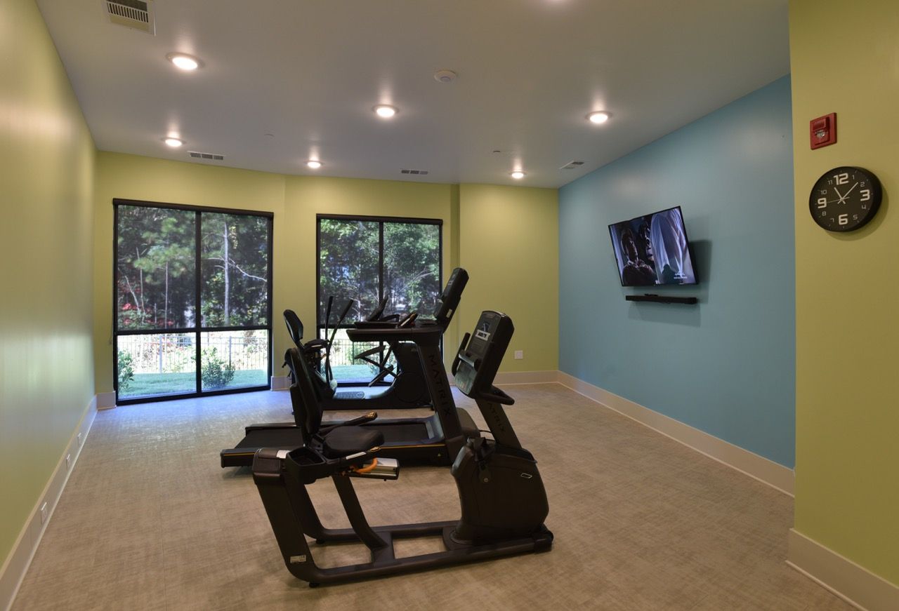 Community fitness center with treadmills, cardio machines, large windows, and a wall-mounted TV.