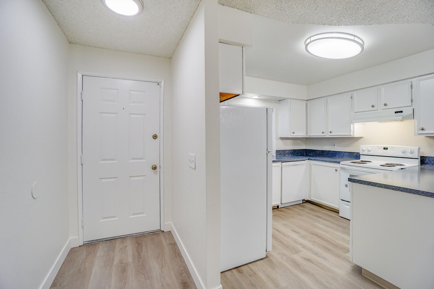 A hallway leading to a kitchen with white cabinets and a refrigerator.