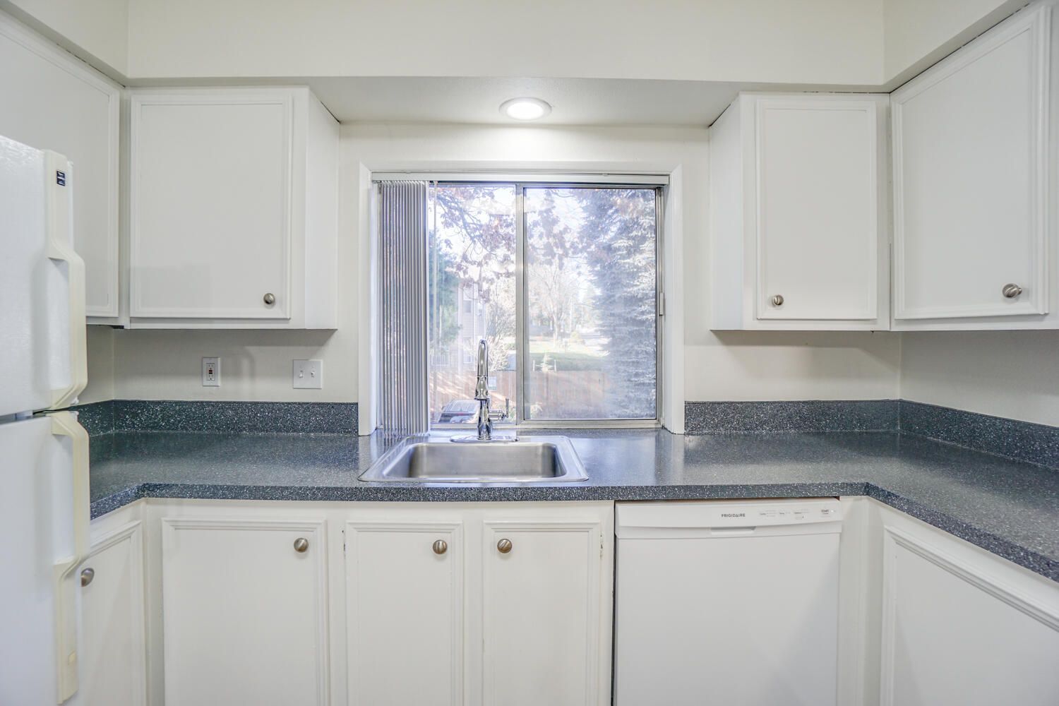 A kitchen with white cabinets , a sink , a refrigerator , and a window.