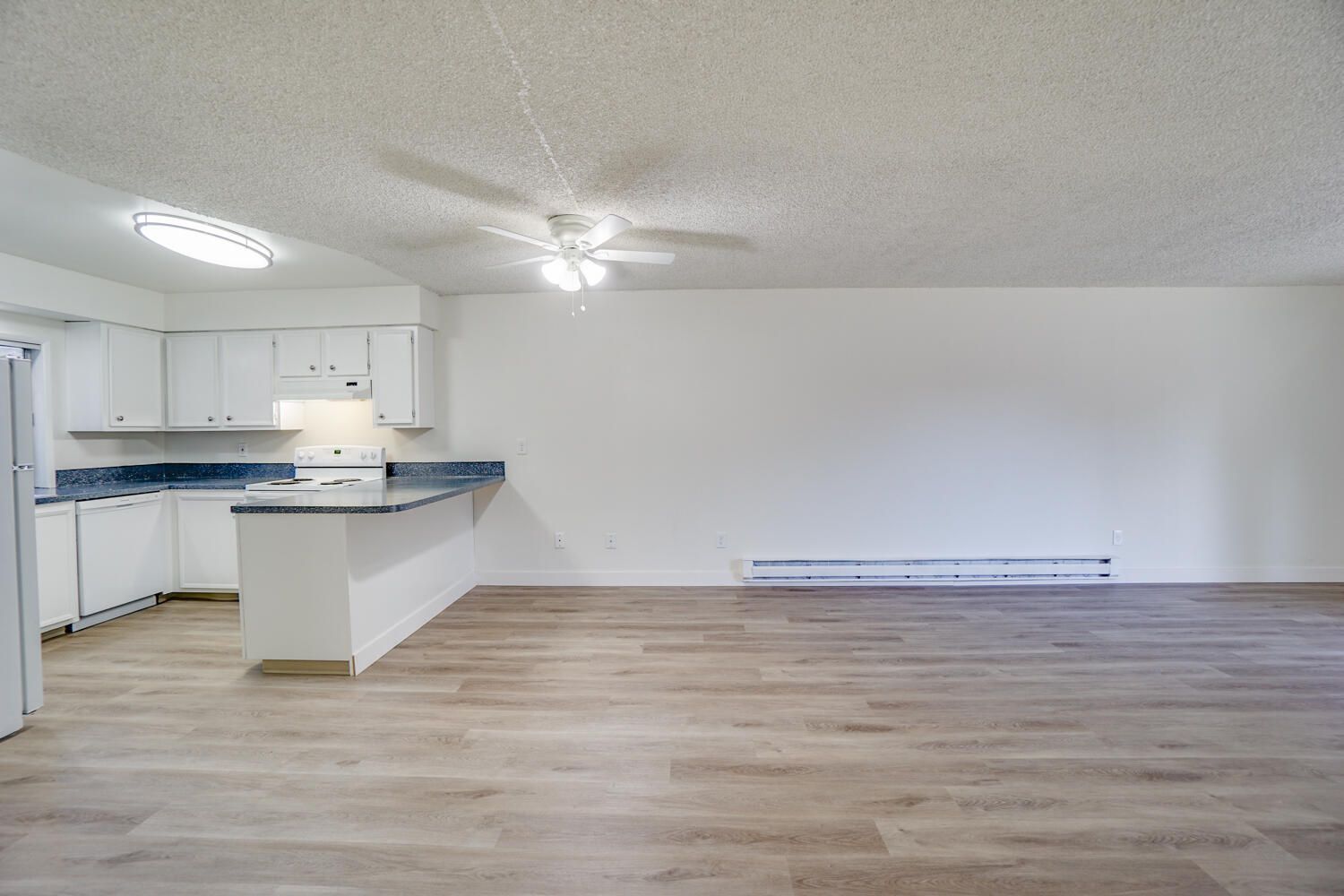 An empty living room with a kitchen and a ceiling fan.
