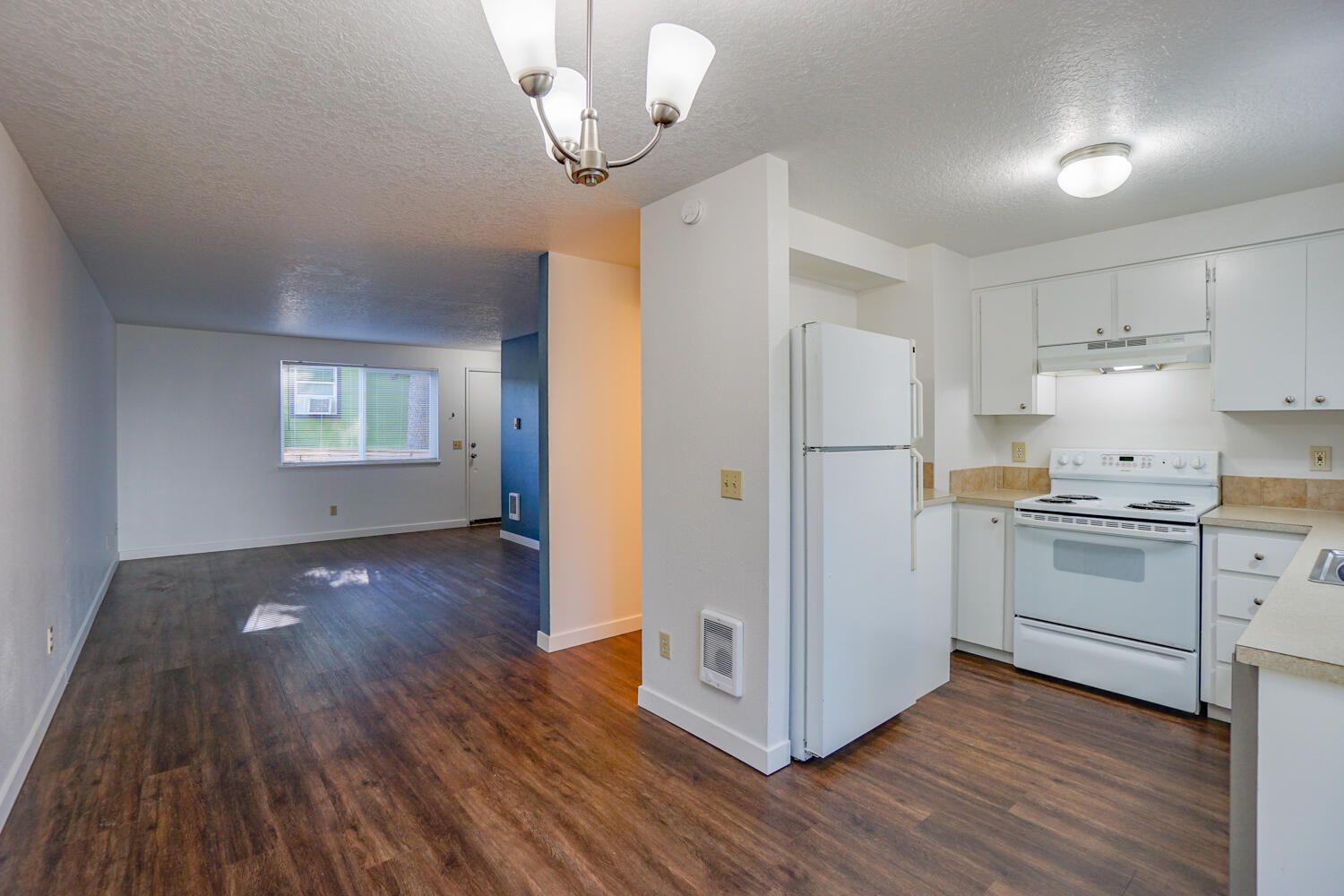 Interior of a white-walled kitchen and living room with hardwood floors and white appliances.