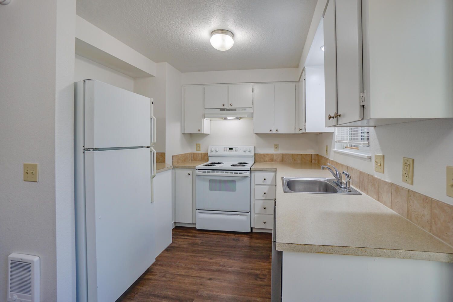 White kitchen with cabinets, appliances, and a sink. Dark brown flooring and beige countertops.
