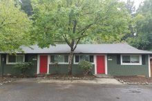 A green house with red doors and white trim is surrounded by trees.