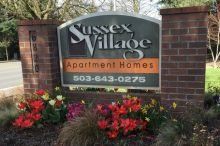 A sign for sussex village apartment homes is surrounded by flowers.