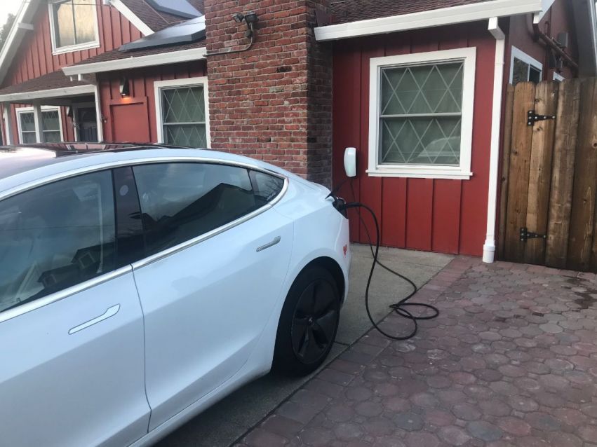White Tesla charging beside a brick house, with a red garage door and driveway cable visible