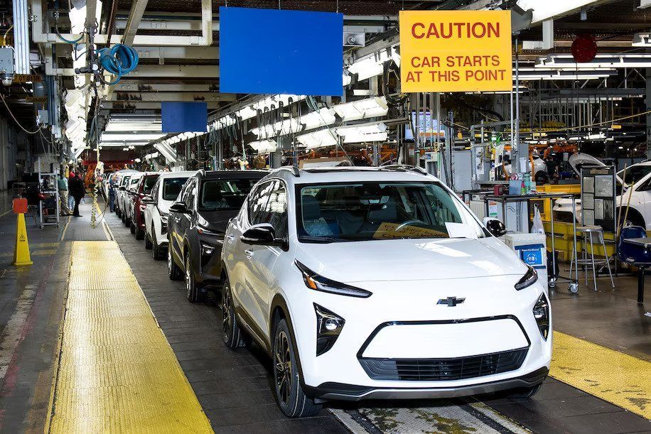 Cars on an assembly line in a factory, with a white SUV in front and a caution sign overhead