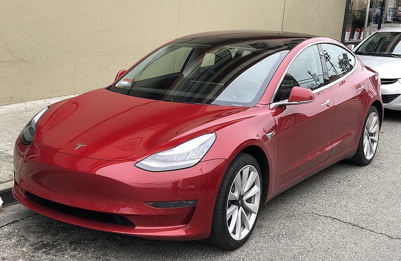 Red Tesla sedan parked on a driveway beside a beige wall, front three-quarter view.