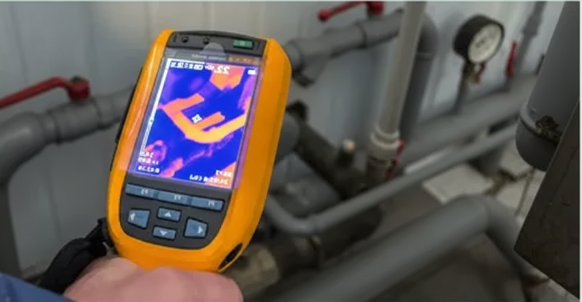 Hand holding a yellow thermal imaging camera pointed at blue pipes.