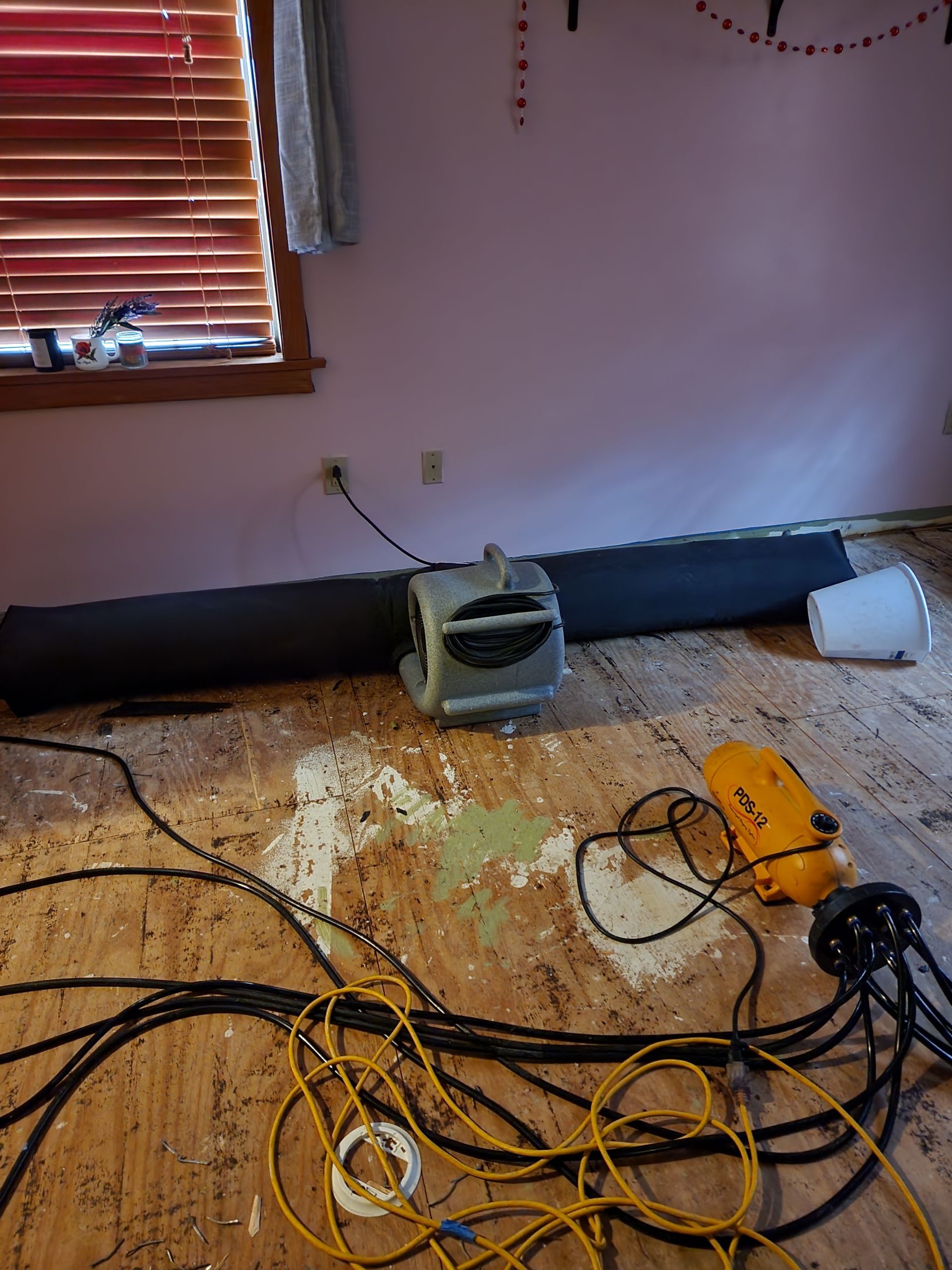 A room with damaged flooring and a fan connected to a long, dark tube. 