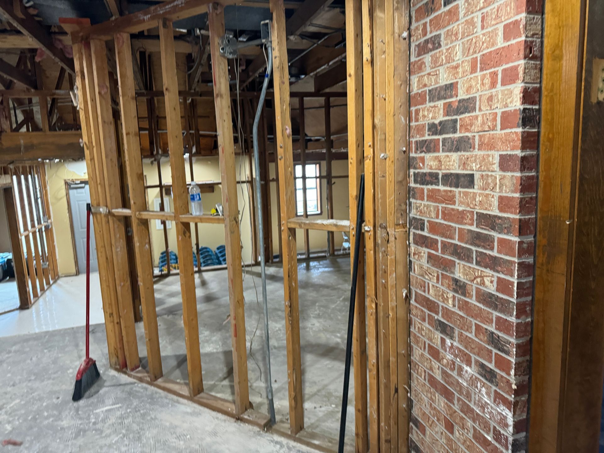 Interior view of a room under renovation with exposed wooden studs and brick wall. A broom rests against a stud.