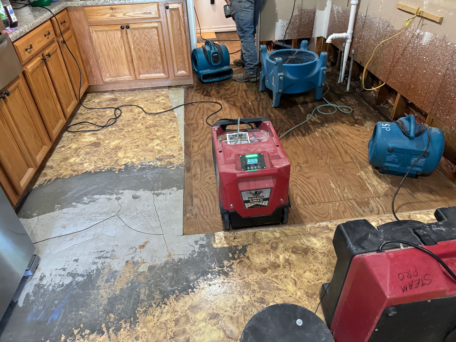Workers use industrial fans and a water extraction machine to dry a flooded kitchen floor with exposed subfloor. 